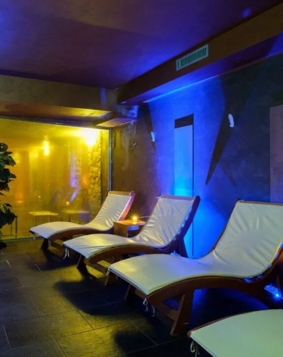 Interno di spa con lettini bianchi, illuminazione soffusa blu e gialla, atmosfera rilassante e pavimento scuro.