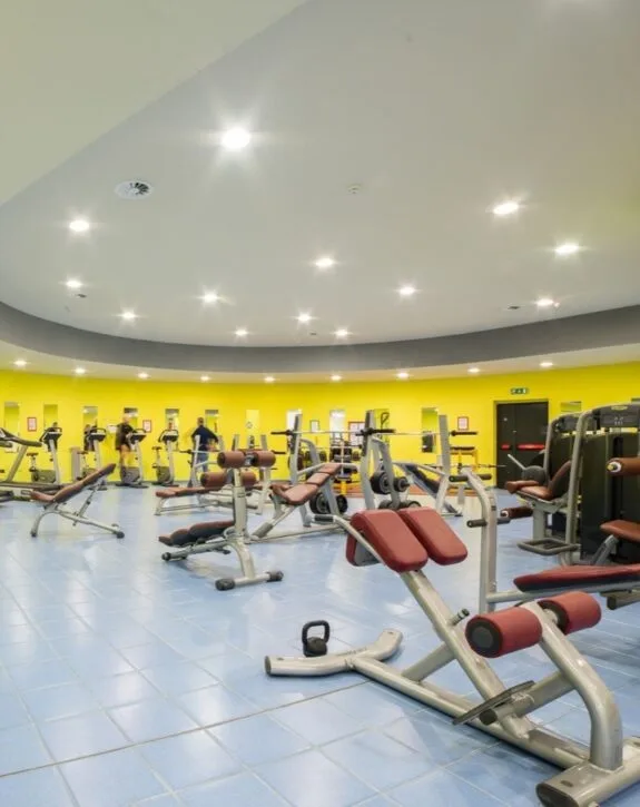 Palestra moderna con attrezzi per allenamento muscolare e cicloergometri su pavimento azzurro e pareti gialle.