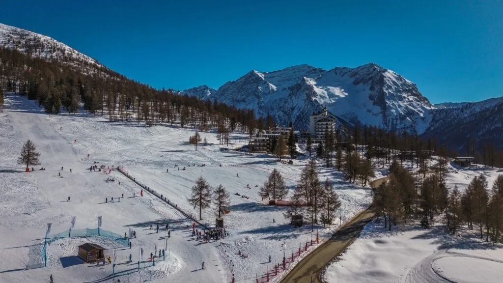 Area sciistica con persone sulla neve, alberi sparsi, edificio e montagne innevate sullo sfondo sotto un cielo azzurro.
