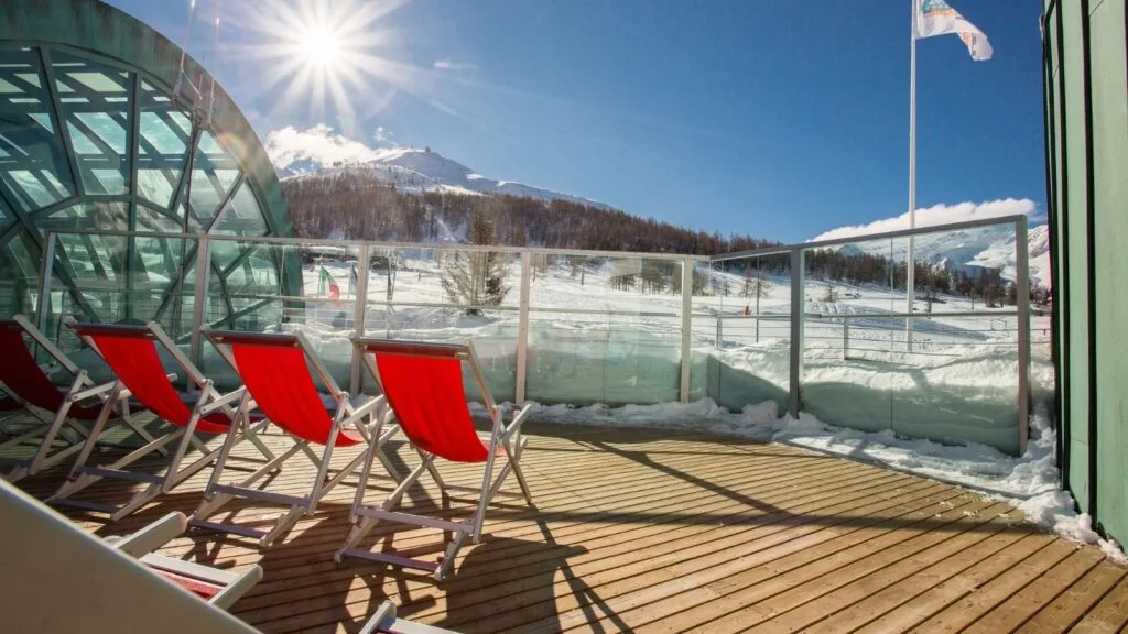 Terrazza con sedie rosse, vista su montagna innevata e cielo soleggiato con sole splendente.