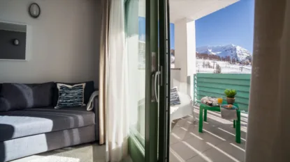 Balcone soleggiato con tavolo verde e colazione, vista su montagne innevate, divano grigio e tenda bianca interna.
