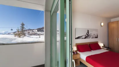 Camera da letto con letto matrimoniale rosso e balcone con vista su paesaggio innevato e sciovie in montagna.