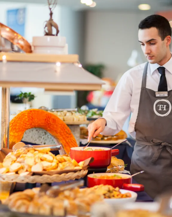 Cameriere in divisa che serve cibo da un buffet con vassoi di pane e decorazione intagliata arancione.