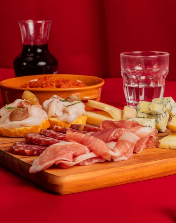 Tagliere con affettati, formaggi misti e crostini, con brocca di vino rosso e bicchiere d'acqua su sfondo rosso.