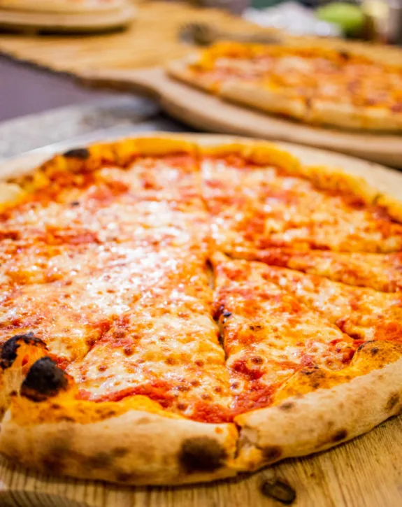 Pizza margherita con mozzarella fusa e salsa di pomodoro su tagliere di legno, sfondo con altra pizza sfocata.