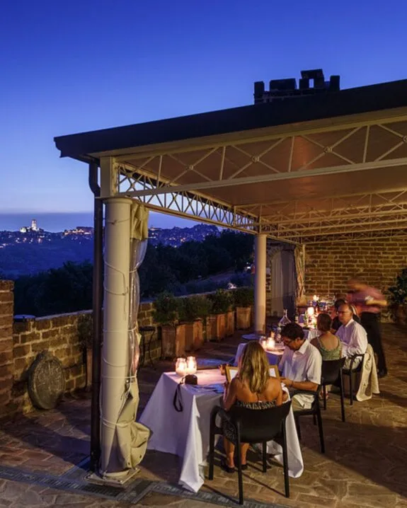 Ristorante all'aperto con clienti che cenano sotto una struttura coperta, vista notturna su colline illuminate in lontananza.