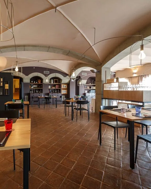 Interno luminoso di ristorante con pavimento in cotto, tavoli in legno, sedie nere e soffitto ad archi con lampade sospese.