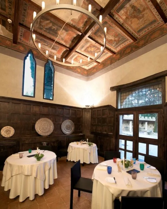 Sala da pranzo elegante con soffitto affrescato, tavoli rotondi con tovaglie bianche e decorazioni floreali.