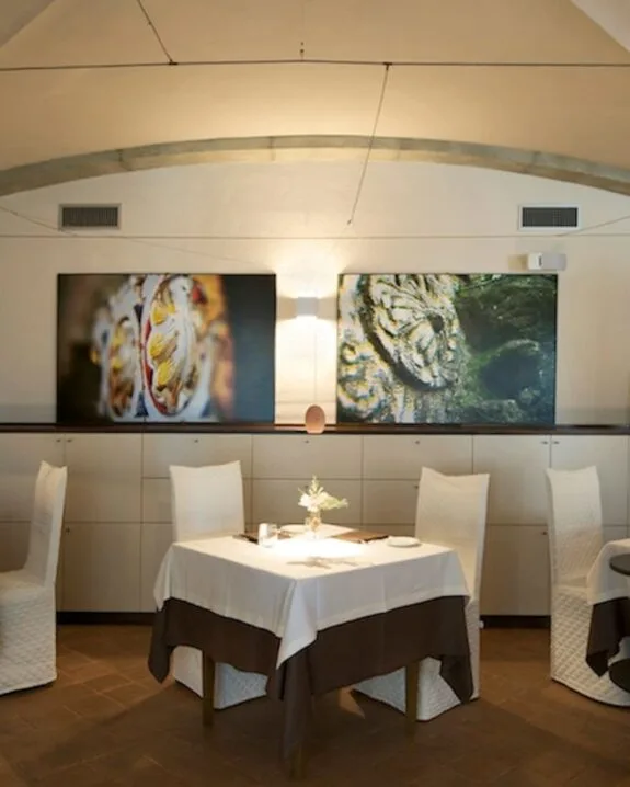 Sala ristorante con tavoli apparecchiati, sedie bianche e due quadri moderni appesi su parete chiara.