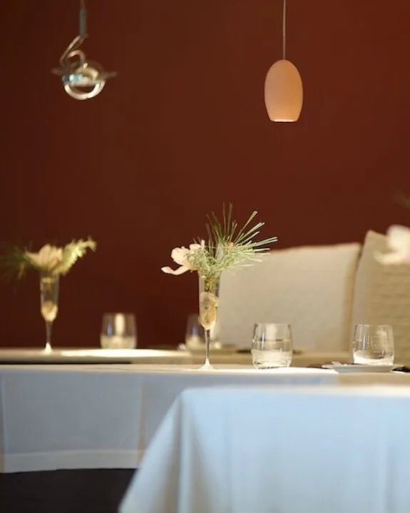 Tavolo apparecchiato con tovaglia bianca, bicchieri, fiori in bicchieri di vetro e luce sospesa su sfondo marrone.
