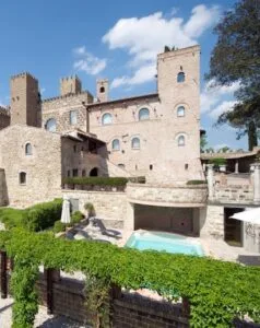 Castello storico in pietra con torre, piscina e giardino verde sotto un cielo azzurro con nuvole.