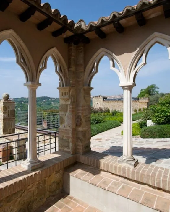 Vista di archi gotici in pietra con colonnine, terrazza con pavimento in mattoni e giardino con mura in lontananza.