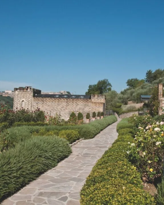 Sentiero in pietra con aiuole verdi e fiori, muro in mattoni e cielo azzurro limpido sullo sfondo.