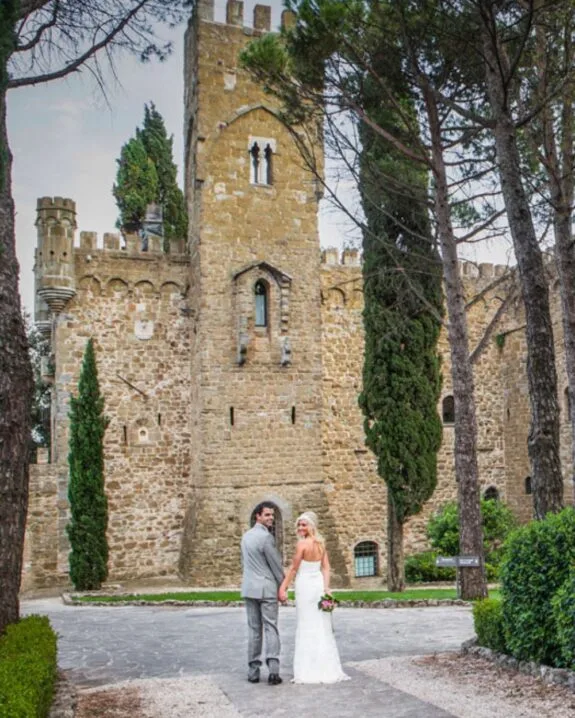 Coppia di sposi che si tengono per mano davanti a un castello medievale circondato da alberi e vegetazione.