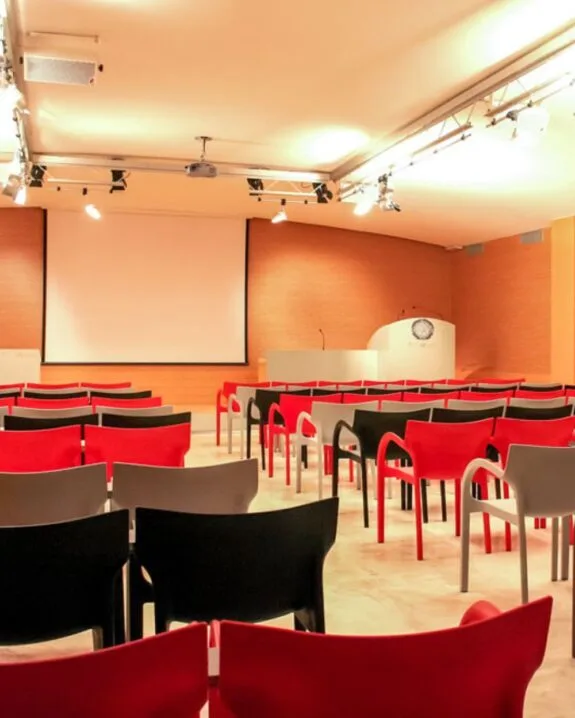 Sala conferenze vuota con sedie rosse, nere e grigie e schermo bianco per presentazioni.