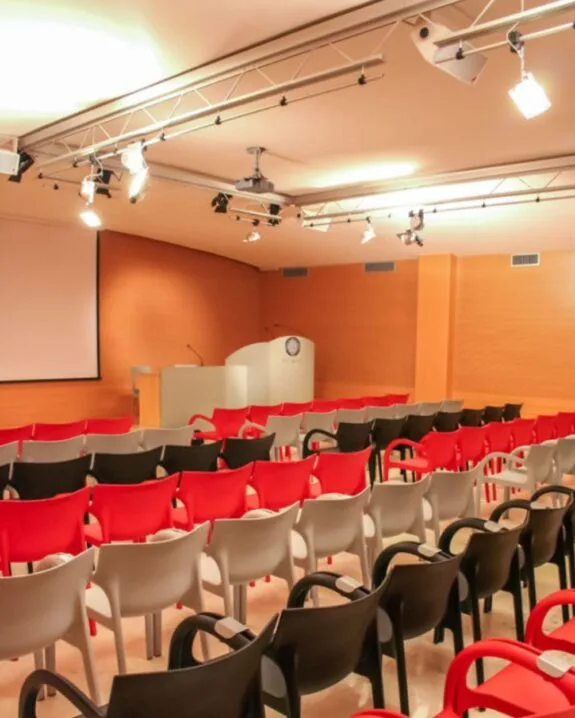 Aula conferenze vuota con file di sedie rosse, nere e bianche di fronte a un podio e uno schermo di proiezione.