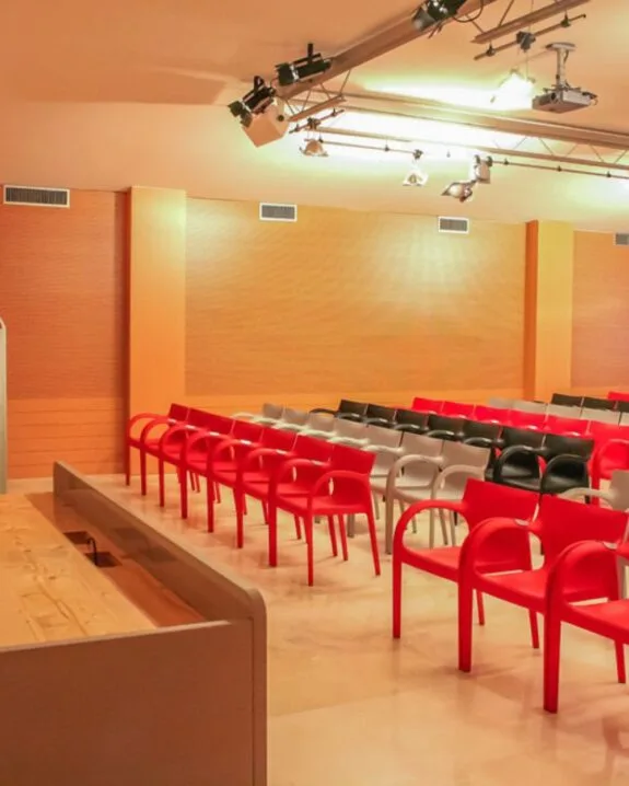 Sala conferenze con sedie rosse, grigie e nere disposte in file su pavimento chiaro.