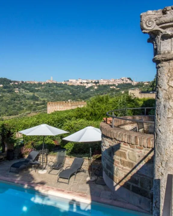 Vista di piscina con lettini e ombrelloni bianchi, muro di pietra e paesaggio collinare verde su cielo azzurro.