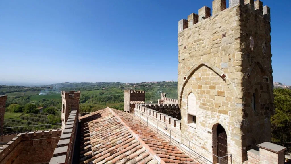 Veduta delle antiche torri di un castello in pietra con tetti in tegole rosse e paesaggio collinare verde sullo sfondo.
