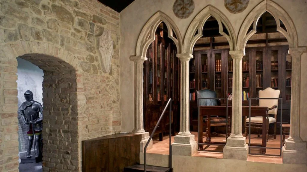 Interno medievale con archi gotici, libreria antica, scrivania, sedie e armatura in pietra e mattoni.