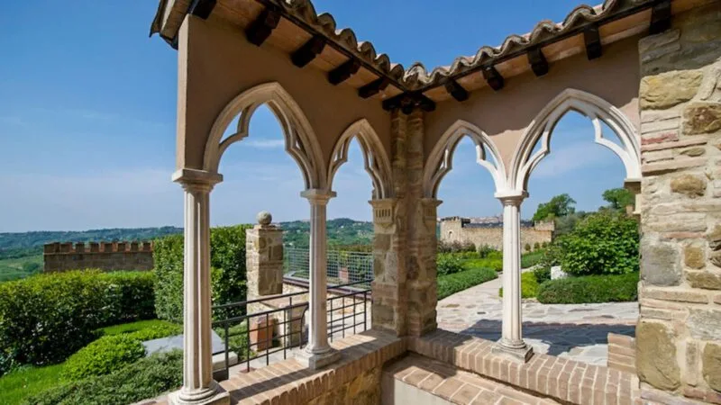 Veduta di un portico con archi gotici in pietra, giardino curato e paesaggio collinare sullo sfondo al sole.