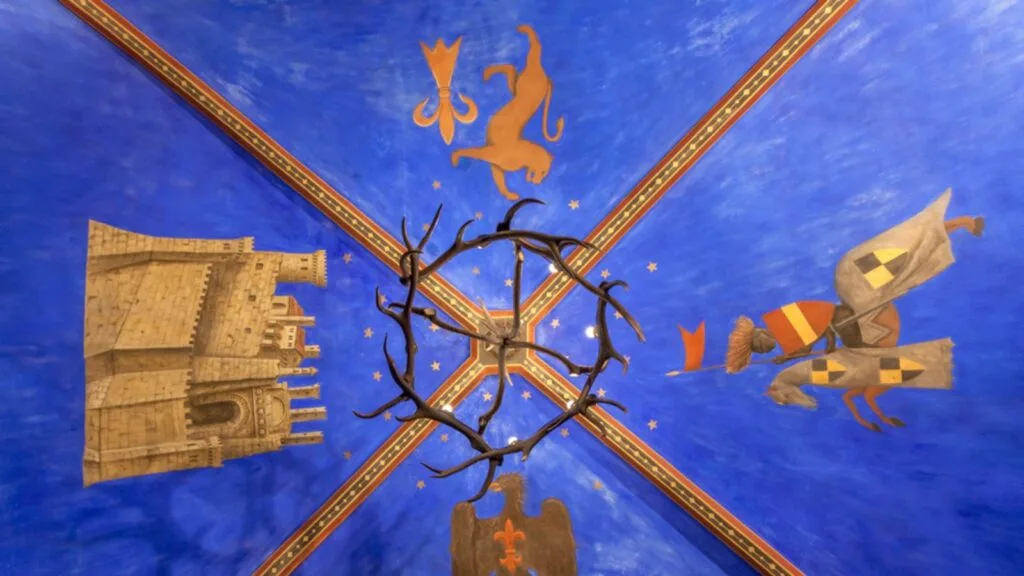 Affresco su soffitto blu con castello, leone, cavaliere a cavallo, aquila e lampadario in ferro battuto.