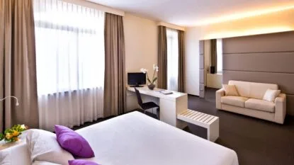 Camera moderna con letto bianco, cuscini viola, scrivania, TV e divano. Grande finestra con tende trasparenti.