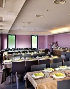Sala ristorante con tavoli preparati per un pasto, tovaglioli gialli e bicchieri, pareti viola e luci sul soffitto.