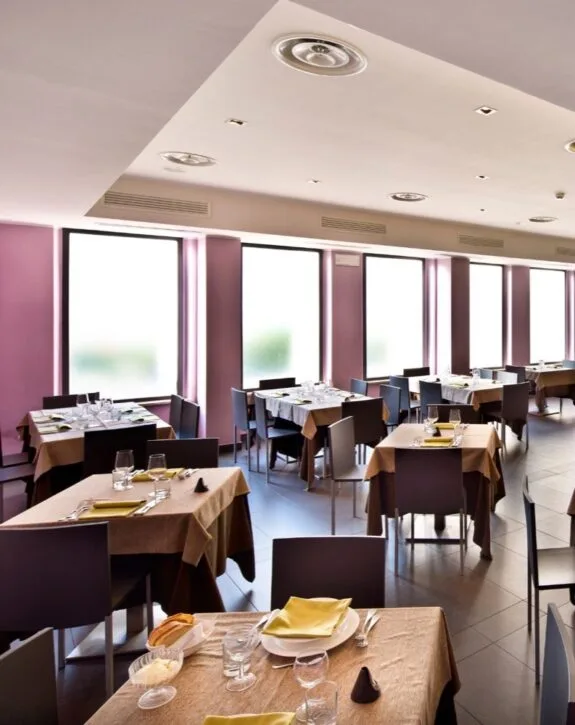Sala ristorante con grandi finestre, tavoli prontos con posate, bicchieri e tovaglioli.