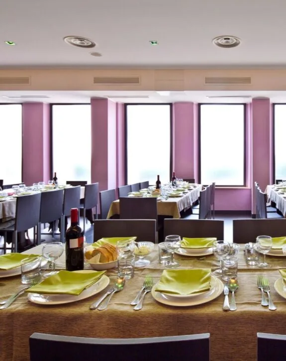 Sala da pranzo in stile moderno con tavoli preparati per un evento, cibo, vino e stoviglie, grandi finestre che lasciano entrare luce naturale.