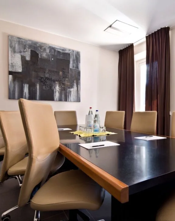 Sala riunioni con tavolo, sedie beige, bottiglie d'acqua e quadri moderni alle pareti.