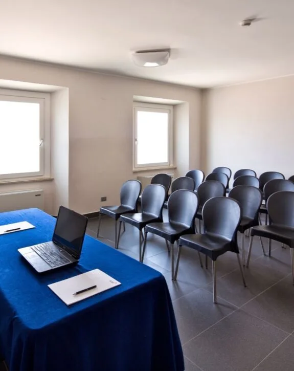 Sala riunioni con sedie nere, tavolo con laptop e quaderno, luce naturale dalle finestre.