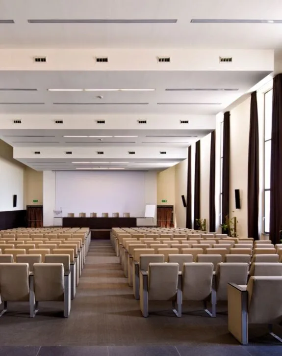 Sale conferenze vuota con sedie beige, grande schermo e pareti con finestre e tende nere.