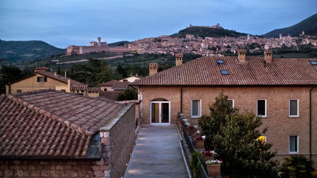 Panorama di Assisi al tramonto, con case in pietra e tetti di tegole visibili in primo piano.