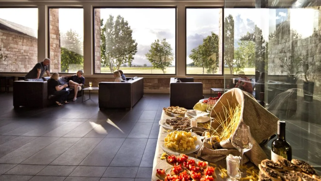 Interno elegante con tavolo di antipasti, vino e persone sedute su divani vicino grandi finestre con vista su alberi.