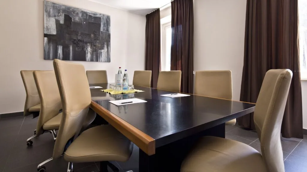 Sala riunioni con tavolo nero, sedie beige e decorazione astratta alle pareti. Due bottiglie e fogli sul tavolo.