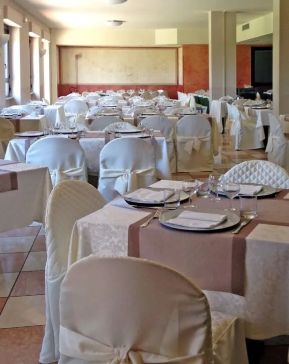 Sala da pranzo elegante con tavoli apparecchiati e sedie coperte da tovaglie e coprisedia bianchi.