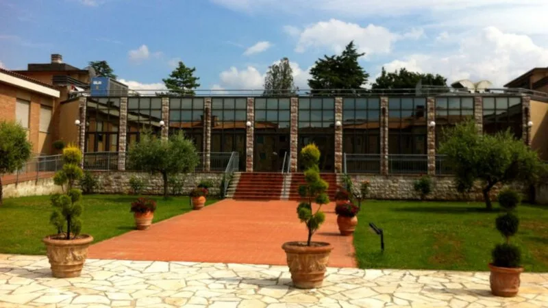 Edificio moderno con facciata di vetro, scala centrale e vasi con piante lungo il vialetto pavimentato.