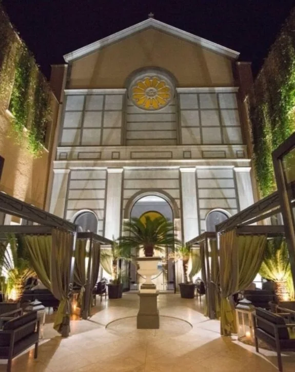Facciata di una chiesa o edificio religioso illuminato di notte, con piante e sedili all'aperto.