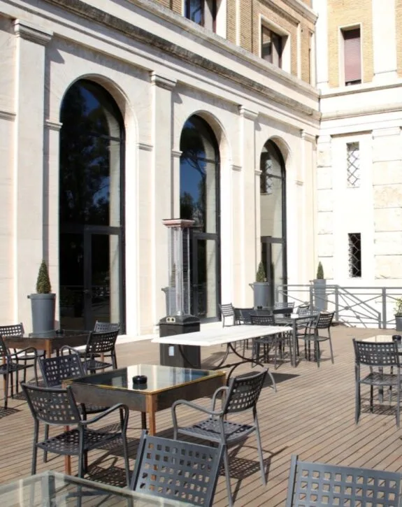 Terrazza esterna con tavoli e sedie in metallo davanti a un edificio con grandi vetrate ad arco.