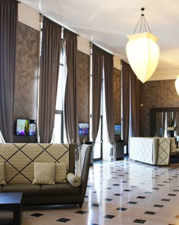 Soggiorno elegante con tende marroni, lampadario decorativo e divani beige con cuscini, pareti decorate e schermi TV.