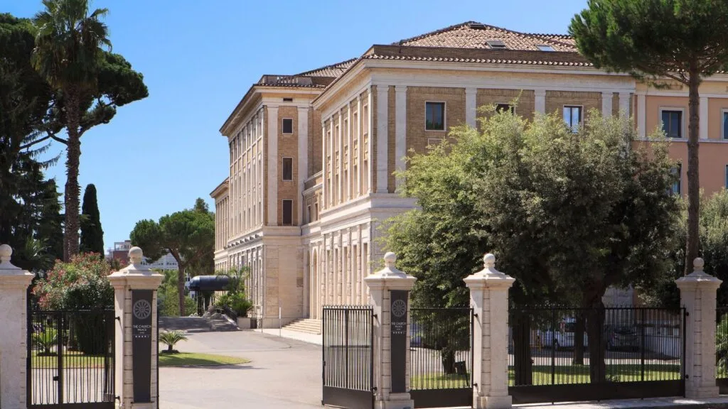 Edificio storico con mura chiare, elementi classici e un cancello nero, circondato da alberi e vegetazione.