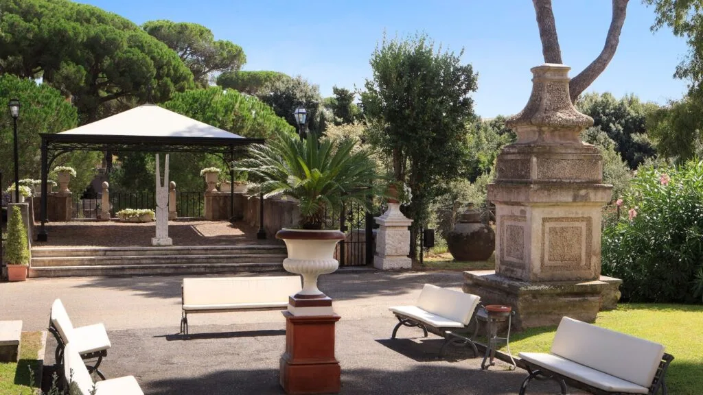 Giardino con panchine, fontana e gazebo sotto alberi verdi e cielo azzurro.