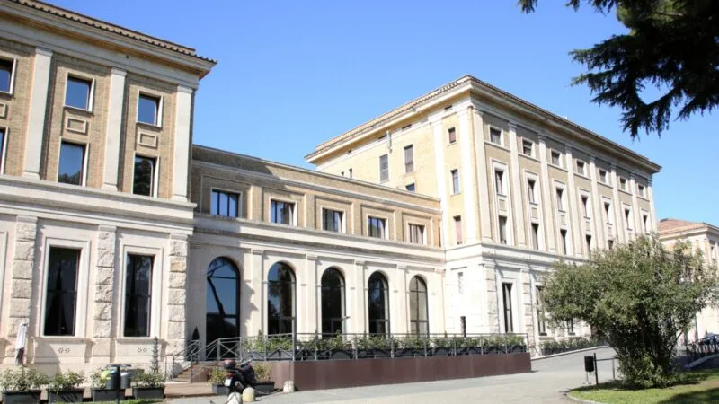 Edificio storico con grandi finestre e archi, immerso nel cielo azzurro e circondato da alberi e panchine.