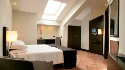 Camera d'hotel luminosa con due letti singoli, pareti chiare, pavimento in cotto e finestra a soffitto.