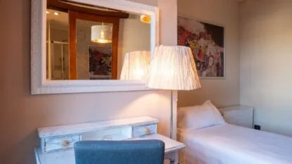Camera con letto singolo, lampada da terra accesa, specchio grande con cornice bianca e scrivania con sedia blu.