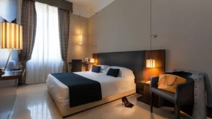 Camera moderna con letto matrimoniale, lampade a sospensione, poltrona con borsa e giacca, e finestra con tende.