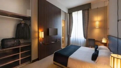 Camera d'hotel moderna con letto matrimoniale, armadio aperto con giacca e borsa, luci calde e finestra con tende.