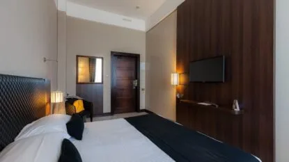 Camera da letto moderna con letto matrimoniale, testiera nera imbottita, TV a parete e poltrona con borsa gialla.