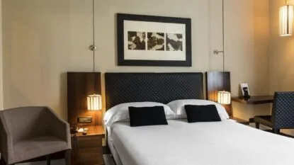 Camera da hotel con letto matrimoniale, testiera nera trapuntata, due cuscini scuri, poltrona grigia e lampade appese ai lati.